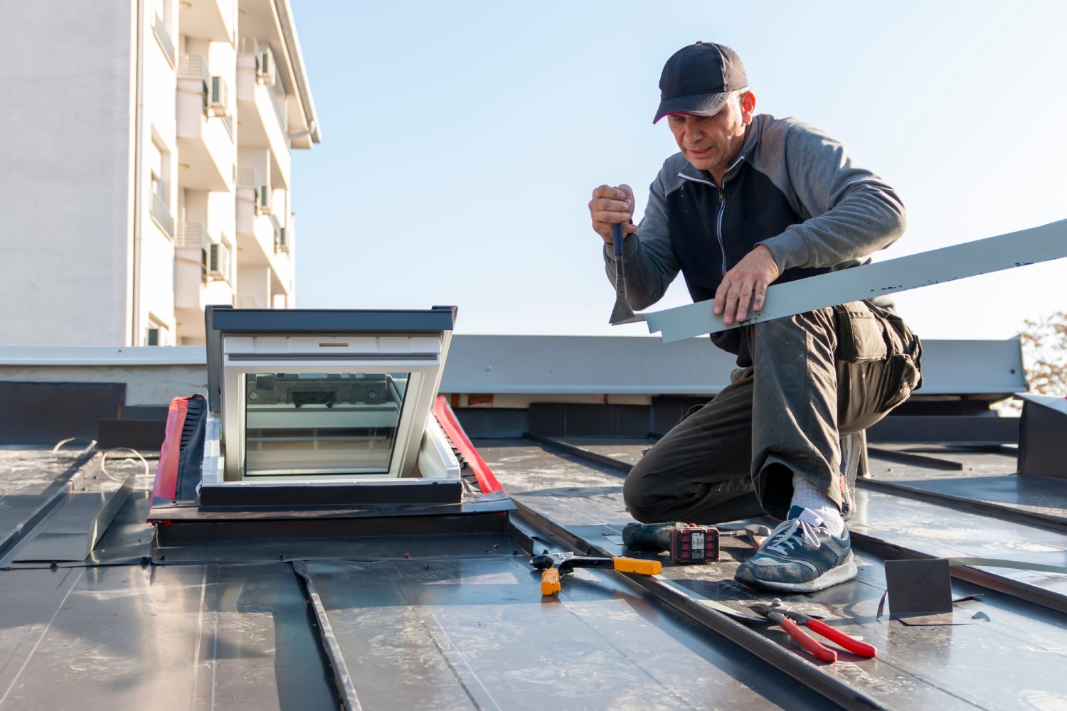 A man kneels on a metal roof, installing a skylight with tools scattered nearby. He wears a cap and jacket, focused on his task. A building is in the background.