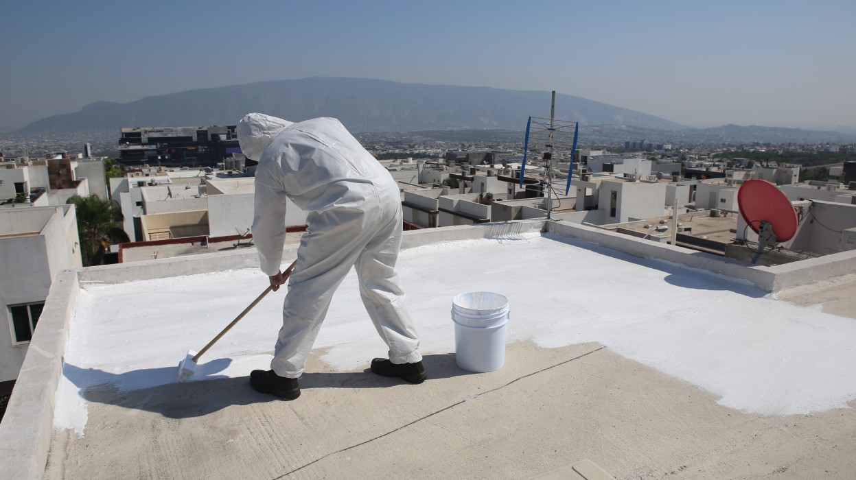 A person in a white suit applies white paint on a rooftop, overlooking a city and mountains under a clear blue sky.